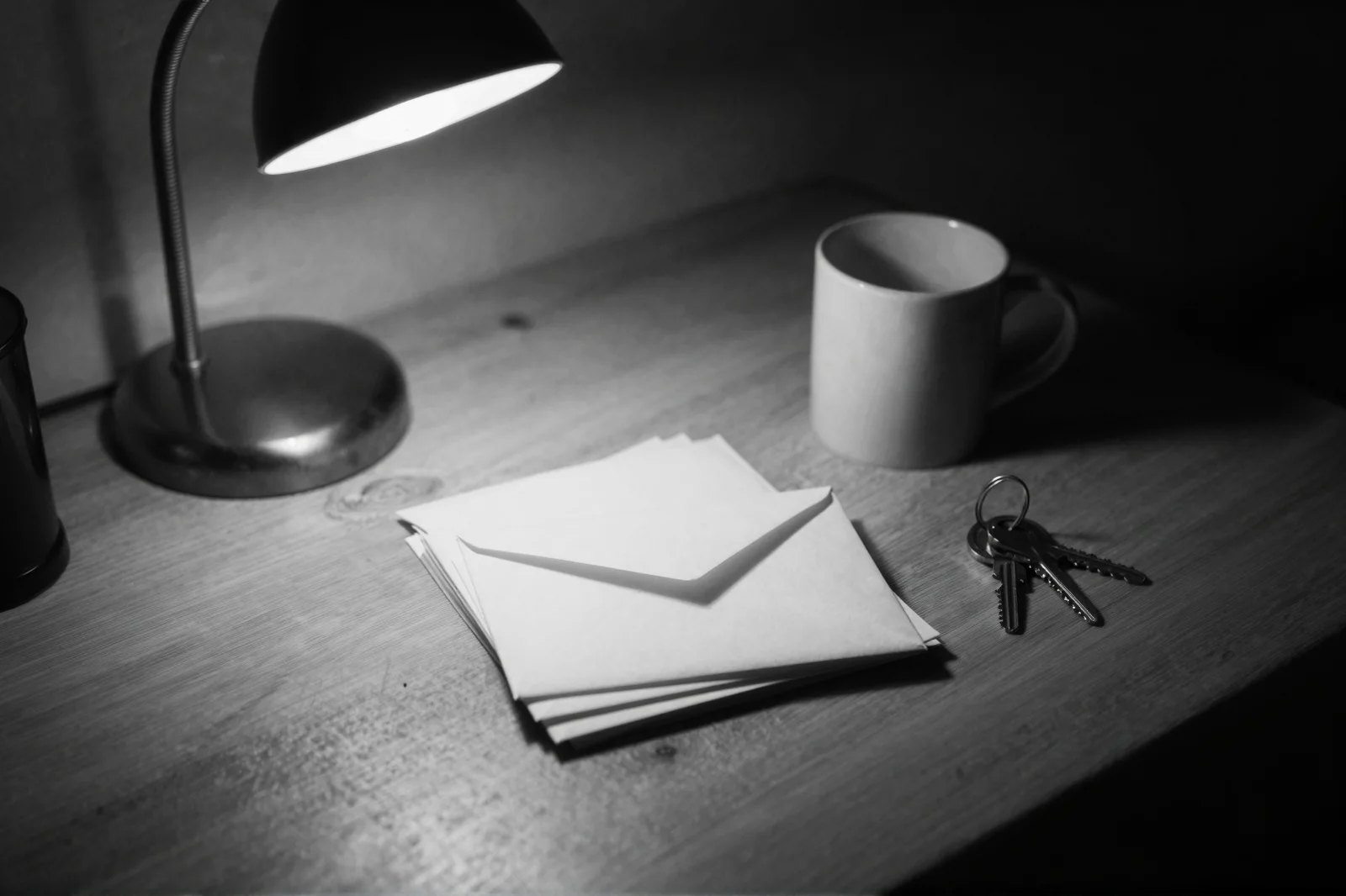 A desk lamp lighting a small stack of envelopes, a mug, and a set of keys on a wooden desk, in black and white.